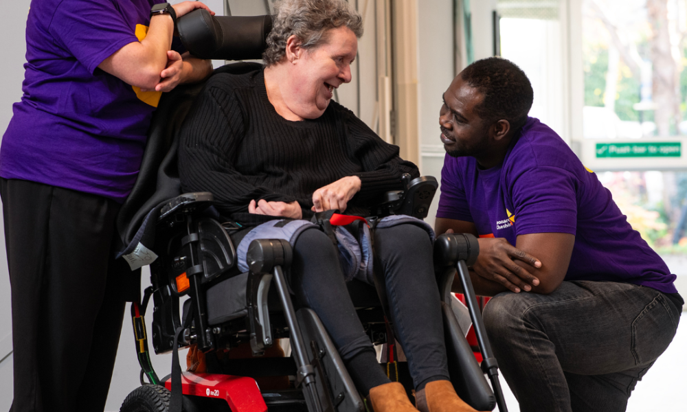 A Caucasian female wheelchair user with two Leonard Cheshire support workers wearing branded t shirts