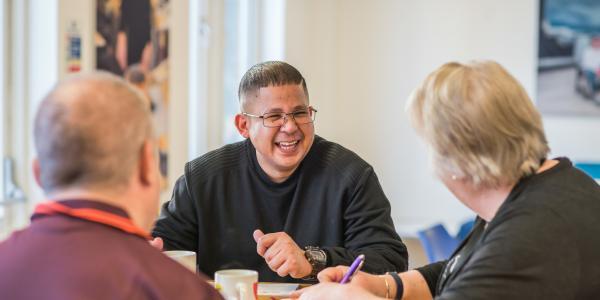 Three people sitting around a table, one is a man with their back to the camera, the other is a woman writing notes and the third is a man wearing glasses and laughing 