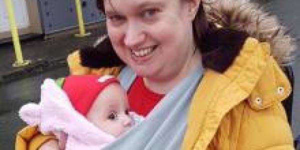 Sarah a Caucasian woman with brown hair wearing a yellow coat and smiling at the camera whilst holding her baby