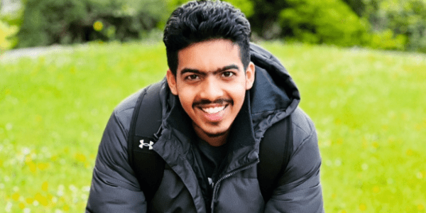 An asian male with black hair and a moustache wearing all black and sat on garden bench with his hands together smiling at the camera with a garden lawn behind him