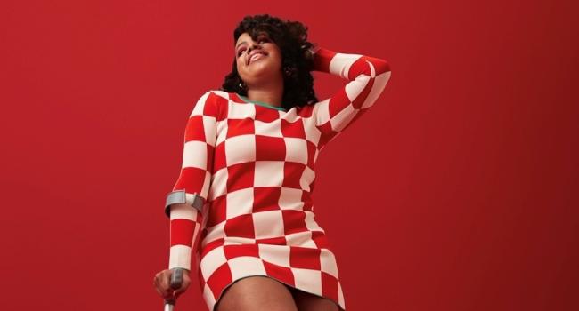 Caprice during a fashion shoot, she is wearing a red and white checked dress, knee high boots and a crutch