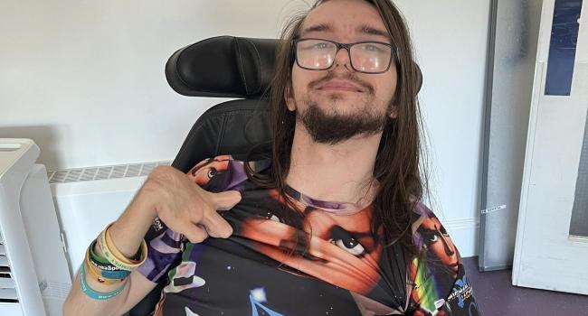 Ryan a caucasian male with long dark hair, a beard and glasses in a powerchair with a mobile phone resting on his wheelchair tray