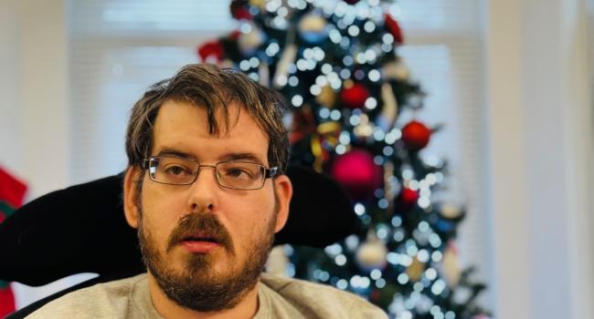 Ashley a caucasian male with short dark hair and a beard wearing glasses and a beige top in front of a Christmas Tree