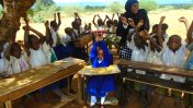 Avina Amos in the school in the outdoor class room, surrounded by other classmates and a teacher with their hands in the air.