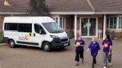 Forecourt of red bricked one level building with a branded minibus parked in it and 3 female care workers wearing purple branded t shirts