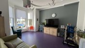 A lounge with smooth purple flooring with a dark green back wall with a tv in front of it on a wooden unit and a green sofa on the other side of the room along with tracking for hoists on the ceiling