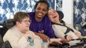 Two caucasian female residents in wheelchairs either side of a female african care worker wearing a purple Leonard Cheshire branded t shirt smiling 