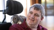 A head shot of a caucasian male resident with grey hair in a power assisted wheelchair with a grey and white bunny sat on his shoulder as he smiles