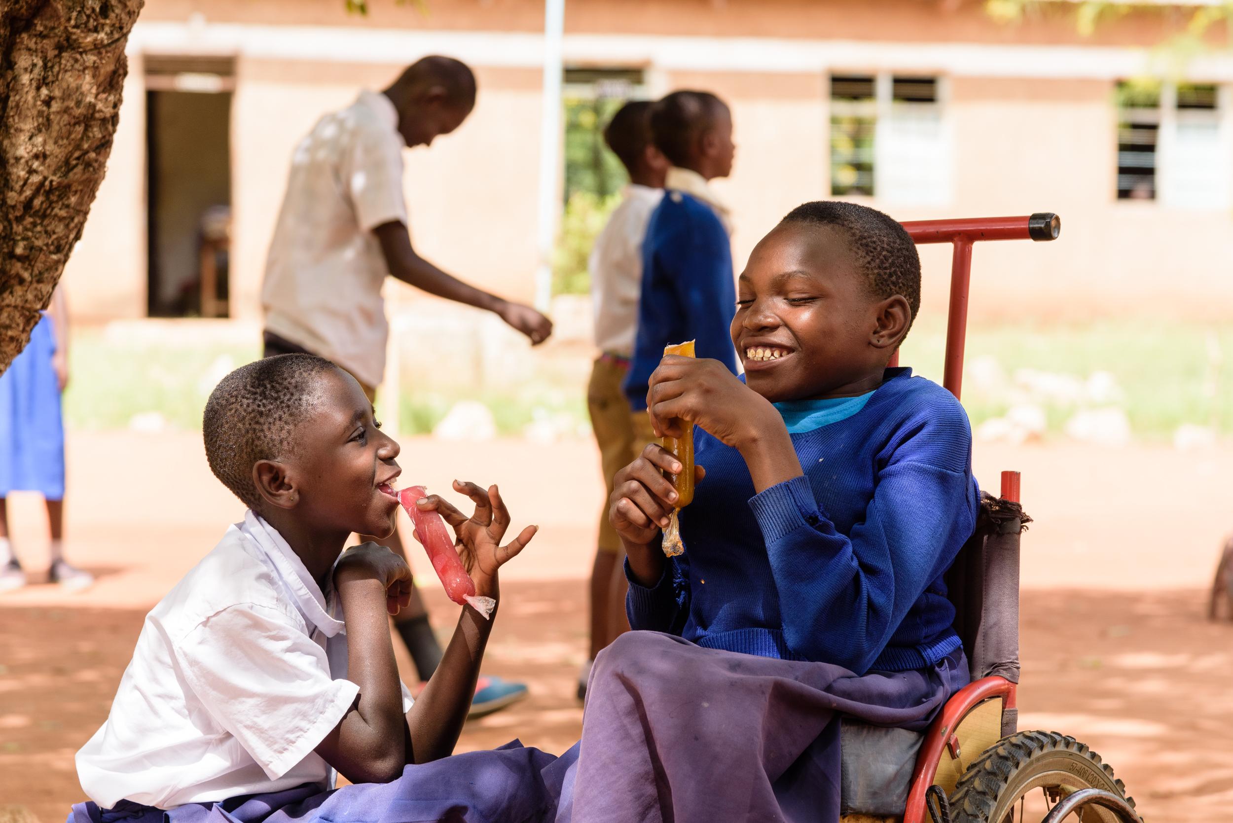 Two young girls sitting outside school eating ice lollies, one is in a wheelchair.