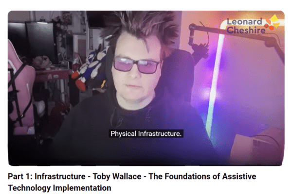 A still shot of Toby a dark haired Caucasian male wearing assistive sunglasses with various lighting elements behind him