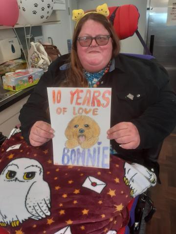 Zoey a caucasian female resident with long brown hair and tinted glasses wearing a black fleece holding up a drawing of Bonnie the dog with the words '10 years of love Bonnie' 