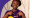 A dark skinned female wearing a branded Leonard Cheshire T shirt and pointing to the logo on it whilst smiling at the camera 