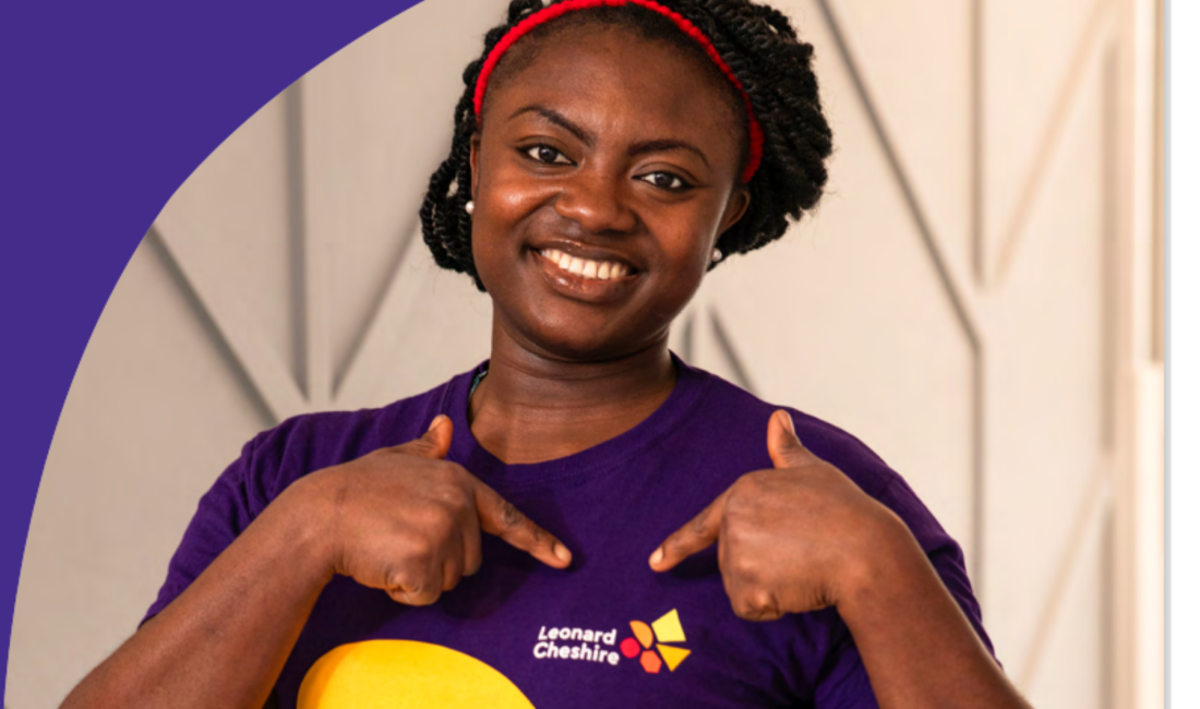 A dark skinned female wearing a branded Leonard Cheshire T shirt and pointing to the logo on it whilst smiling at the camera