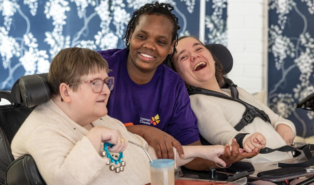Two caucasian female residents in wheelchairs either side of a female african care worker wearing a purple Leonard Cheshire branded t shirt smiling 
