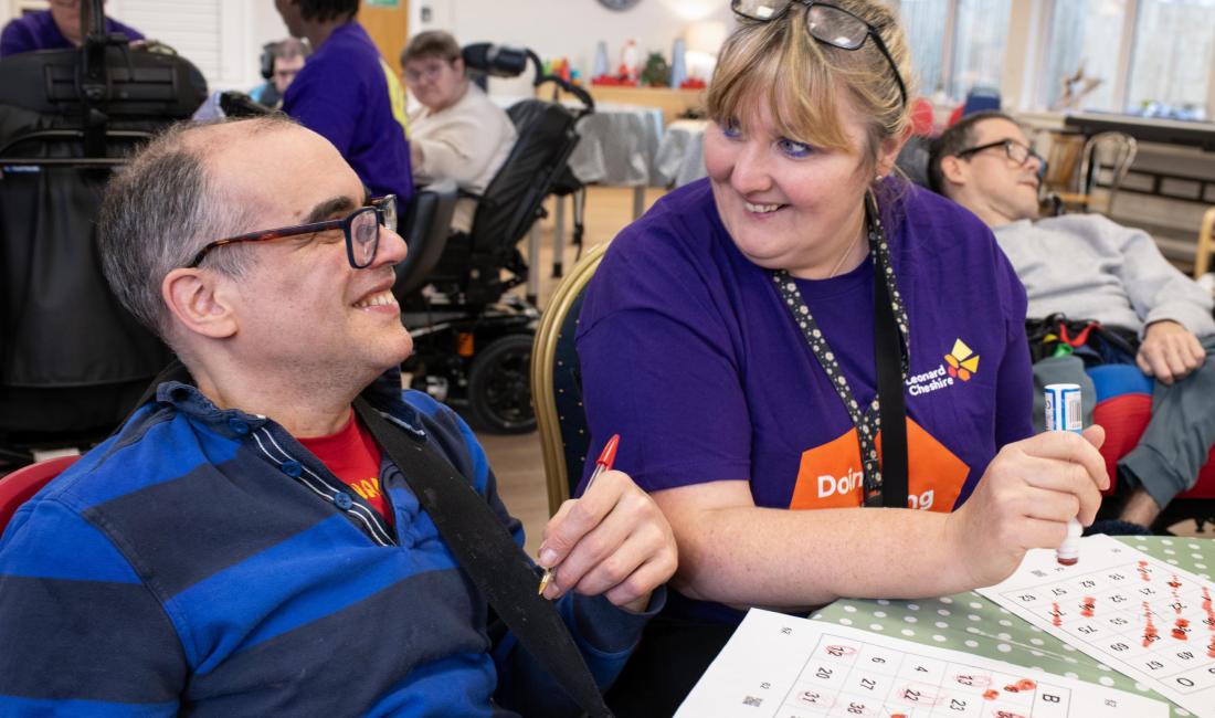 A caucasian male resident with glasses and a blue striped rugby shirt smiling at a caucasian female care worker wearing a purple t shirt both holding dabbers at a bingo game